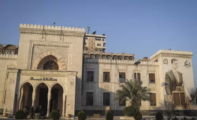 Opposition fighters, left, stand at the entrance of the provincial government office, where an image of Syrian President Bashar Assad is riddled with bullets on the facade, in the aftermath of the opposition's takeover of Hama, Syria, on Friday, Dec. 6, 2024. (AP Photo/Omar Albam)