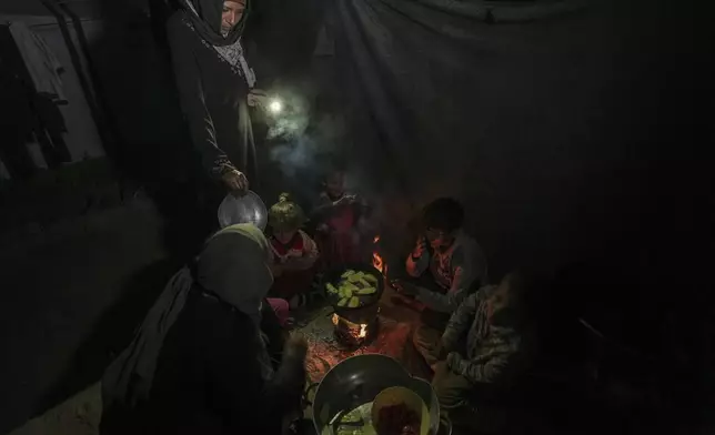 Reda Abu Zarada, left, and her daughter, Amani, standing, displaced from Jabaliya in northern Gaza, feed their children and grandchildren with fried zucchini made over a fire made of paper and cardboard scraps outside their tent in a camp in Khan Younis, Gaza Strip, Thursday, Dec. 19, 2024. (AP Photo/Abdel Kareem Hana)