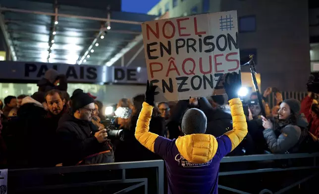 A man with a placard reading "Christmas in prison, Easter in the slammer" walks past the media as they wait outside the courthouse of Avignon during the trial of Dominique Pelicot, in Avignon, southern France, Thursday, Dec. 19, 2024. (AP Photo/Lewis Joly)