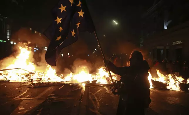 A demonstrator holds a European Union flag standing next to a burning barricade during a rally to protests against the government's decision to suspend negotiations on joining the EU in Tbilisi, Georgia, early Tuesday, Dec. 3, 2024. (AP Photo/Zurab Tsertsvadze)