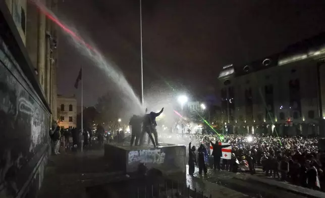 Demonstrators stand atop of a tomb under running water from a water cannon rallying outside the parliament's building to continue protests against the government's decision to suspend negotiations on joining the European Union in Tbilisi, Georgia, on Monday, Dec. 2, 2024. (AP Photo/Zurab Tsertsvadze)