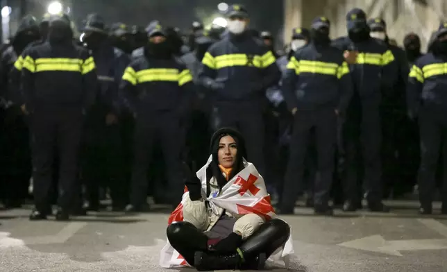 A demonstrator draped in a Georgian national flag sits in front of police rallying outside the parliament's building to continue protests against the government's decision to suspend negotiations on joining the European Union in Tbilisi, Georgia, on Monday, Dec. 2, 2024. (AP Photo/Zurab Tsertsvadze)