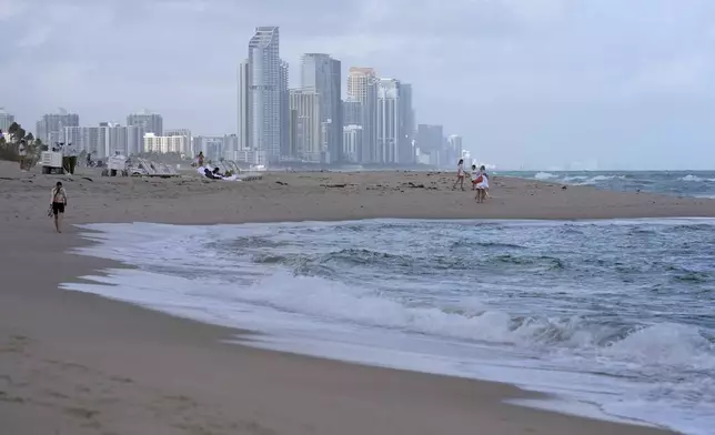 People walk along the beach in Surfside, Fla., near the skyline of Sunny Isles Beach, Tuesday, Dec. 17, 2024. (AP Photo/Lynne Sladky)