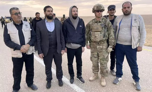 This image provided by Mouaz Moustafa shows Travis Timmerman, center, standing with a member of the U.S. military and representatives of Syria's transitional government and opposition activists near the Syria-Jordanian border Friday, Dec. 13, 2024. U.S. forces flew Timmerman out of Syria Friday after rebels freed him following seven months in a Syrian government prison. (Mouaz Moustafa via AP)