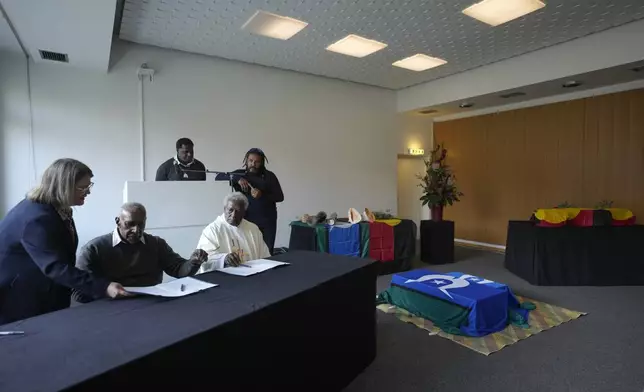 From left: The Australien Ambassador to Germany Natasha Smith and the elders from the Ugar Island, Tomson Stephen, center and Father Daniel Stephen, right, sign documents next to the coffins, during a ceremony for the return of Aboriginal Australians ancestors to their relatives and communities at the Ethnological Museum in Berlin, Germany, Thursday, Dec. 5, 2024. (AP Photo/Markus Schreiber)