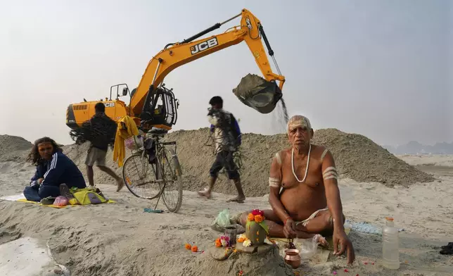 A Hindu holy man prays sitting next to an earthmover moving sand on the banks of the Ganges river, in preparation for the upcoming Maha Kumbh Mela, one of the world's largest religious festivals, in Prayagraj, India, Friday, Nov. 29, 2024. (AP Photo/Rajesh Kumar Singh)