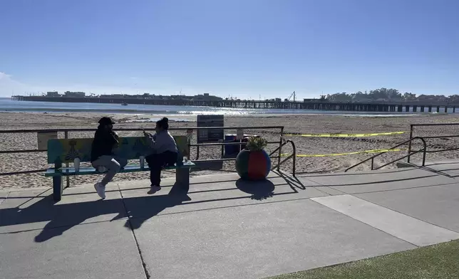 People sit on a bench along the beach in Santa Cruz, Calif. after authorities cleared the main beach, taping off entrances on Thursday, Dec. 5, 2024. (AP Photo/Martha Mendoza)