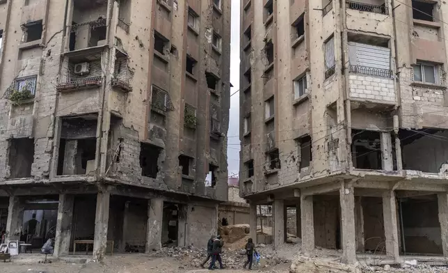 People walk between buildings that were damaged by airstrikes during the Assad rule in Douma, on the outskirts of Damascus, Syria, Sunday, Dec. 29, 2024 (AP Photo/Mosa'ab Elshamy)