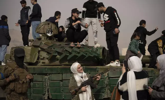 A woman holding a rifle borrowed from a Syrian opposition fighter poses for a picture, next to a government forces tank that was left on a street, at the Umayyad Square in Damascus, Syria, Wednesday, Dec. 11, 2024. (AP Photo/Leo Correa)