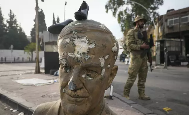 An opposition fighter in military uniform stands next to a broken bust of the late Syrian President Hafez Assad, with a pair of shoes placed on top, at a checkpoint in Damascus, Syria, Wednesday, Dec. 11, 2024. (AP Photo/Hussein Malla)