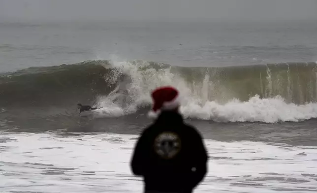 Jeff Parker, wearing a Santa Claus hat, watches as a surfer rides a wave in Seal Beach, Calif., Tuesday, Dec. 24, 2024. (AP Photo/Jae C. Hong)