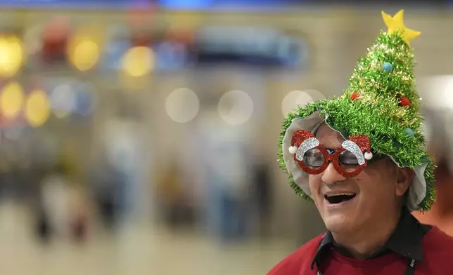 Cesar Davila, who works for the American Airlines partner providing wheelchair assistance, wears a Christmas tree hat and festive glasses as he waits for a traveler to help inside the American Airlines terminal at Miami International Airport, on Christmas Eve, Tuesday, Dec. 24, 2024, in Miami. (AP Photo/Rebecca Blackwell)