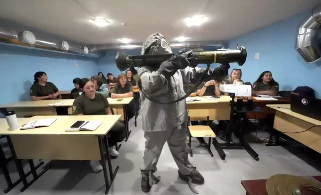 A Ukrainian serviceman in protective suit demonstrates grenade launcher to women during a training course for national resistance for local population in Kharkiv region, Ukraine, Friday, Sept.13, 2024. (AP Photo/Andrii Marienko)