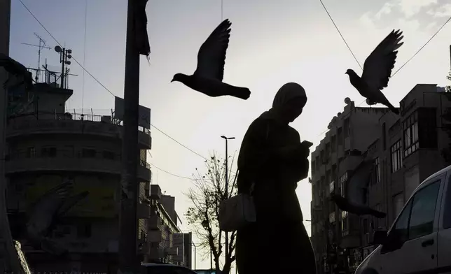 A woman walks amid pigeons flying in a square in the West Bank city of Ramallah, Friday, Dec. 27, 2024. (AP Photo/Matias Delacroix)