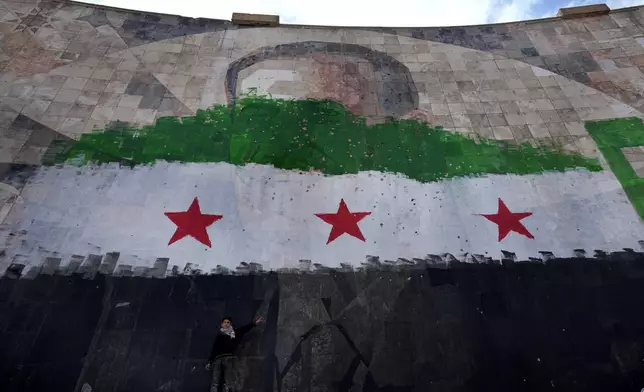 A Syrian boy stands under a giant portrait of the late Syrian President Hafez Assad painted over with the colors of the "revolutionary" flag, in Damascus, Syria, Thursday, Dec. 26, 2024. (AP Photo/Hussein Malla)