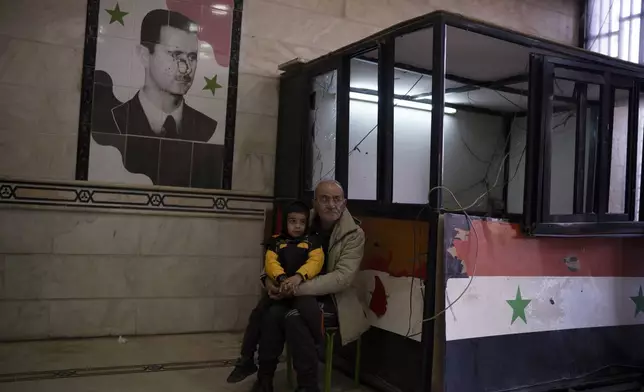 A man sits with a child next to a damaged image depicting the ousted Syrian President Bashar Assad, at the entrance of the Police headquarters, in Homs, Syria, Thursday, Dec. 26, 2024. (AP Photo/Leo Correa)