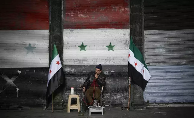 A man smokes a water pipe next to two Syrian "revolutionary" flags in front of a door still adorned with the official flag of the ousted government, near the Umayyad Mosque ahead of Friday prayers in Damascus, Syria, Friday, Dec. 20, 2024. (AP Photo/Leo Correa)