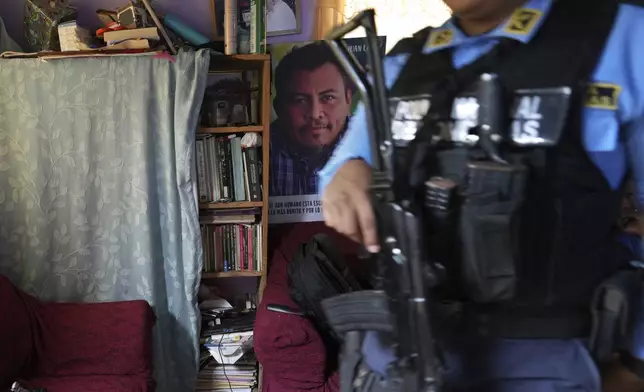 A poster of slain environmentalist Juan López hangs in his home as a state-assigned bodyguard watches over the López family, in Tocoa, Honduras, Friday, Dec. 6, 2024. (AP Photo/Moises Castillo)