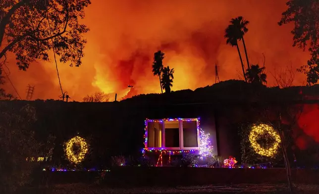 A helicopter drops water on the Palisades Fire behind a home with Christmas lights in Mandeville Canyon, Friday, Jan. 10, 2025, in Los Angeles. (AP Photo/Ethan Swope)