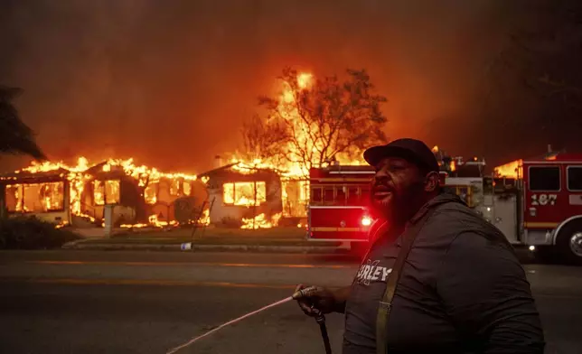 A resident sprays their property with a garden hose as the Eaton Fire engulfs structures across the street, Wednesday, Jan. 8, 2025 in Altadena, Calif. (AP Photo/Ethan Swope)