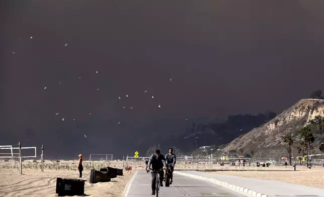 Bike riders make their way along the coast as large dark plume of smoke passes over the beach from a wildfire from Pacific Palisades, from Santa Monica, Calif., on Tuesday, Jan. 7, 2025. (AP Photo/Richard Vogel)