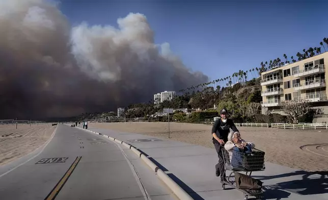 Jerome Krausse pushes his mother-in-law in a shopping cart as they evacuate from their home in the Pacific Palisades after a wildfire swept through their neighborhood in Santa Monica, Calif., on Tuesday, Jan. 7, 2025. (AP Photo/Richard Vogel)