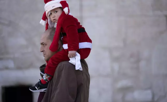 Anton Nakle carries his three year-old son, David, dressed in a tiny Santa Claus suit, into the Church of the Nativity, where Christians believe Jesus Christ was born, in the West Bank city of Bethlehem, on Christmas Eve for Orthodox Christians, Monday, Jan. 6, 2025. (AP Photo/Maya Alleruzzo)