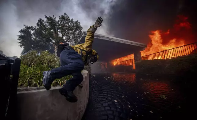 A firefighter jumps over a fence while fighting the Palisades Fire in the Pacific Palisades neighborhood of Los Angeles, Tuesday, Jan. 7, 2025. (AP Photo/Ethan Swope)