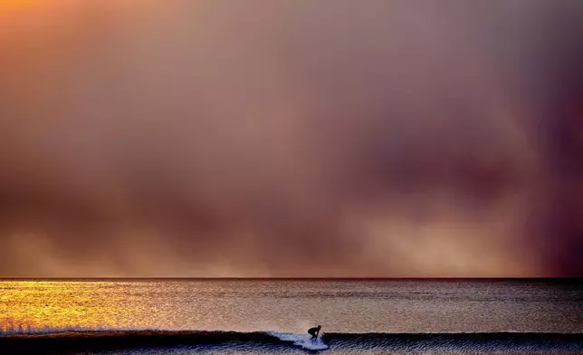 A surfer takes off on a wave in Santa Monica, Calif., during sunset under a blackened sky from the Palisades fire in the Pacific Palisades on Tuesday, Jan. 7, 2025. (AP Photo/Richard Vogel)