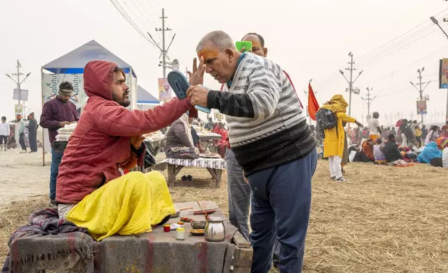 A Hindu devotee looks into a mirror as he gets his forehead smeared by a holy man at the confluence of the Ganges, the Yamuna and the mythical Saraswati rivers, a day before the official beginning of the 45-day-long Maha Kumbh festival, in Prayagraj, India, Sunday, Jan. 12, 2025. (AP Photo/Ashwini Bhatia)