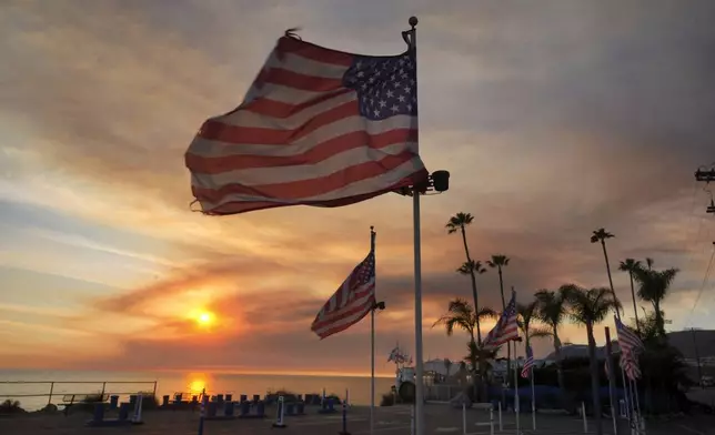 FILE - Flags fly under heavy winds before sunset as a plume of smoke from the Franklin Fire rises over the ocean Tuesday, Dec. 10, 2024, in Malibu, Calif. (AP Photo/Damian Dovarganes,File)