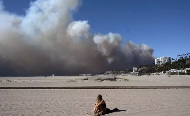 A lone sunbather sits and watches a large plume of smoke from a wildfire rise over the Pacific Palisades, in Santa Monica, Calif., on Tuesday, Jan. 7, 2025. (AP Photo/Richard Vogel)