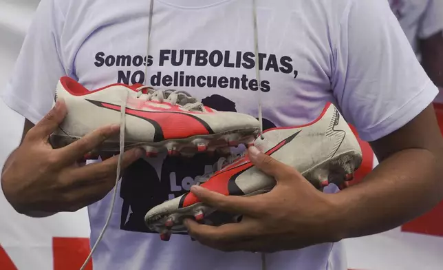 A protester holds soccer cleats and wears a shirt that reads in Spanish "We're soccer players, not delinquents" outside the court holding a hearing for the soldiers accused of being connected to the disappearance of four children in Guayaquil, Ecuador, Tuesday, Dec. 31, 2024. The children disappeared after playing soccer. (AP Photo/Cesar Munoz)