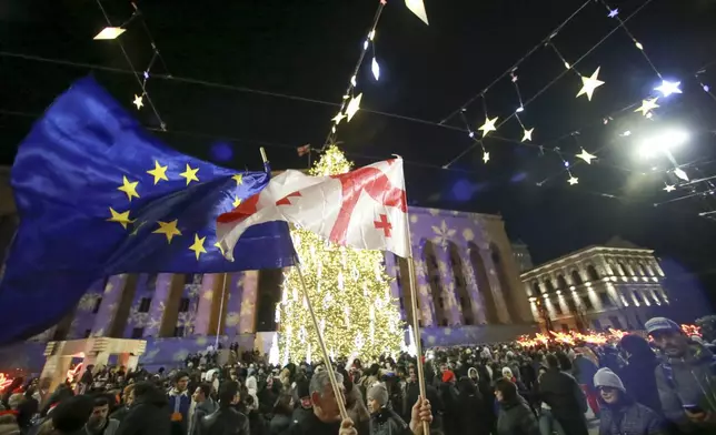 People with a Georgian national and an EU flags gather in a street decorated for Christmas and the New Year festivities on New Year's Eve outside of the Georgian parliament, in Tbilisi, Georgia, on Tuesday, Dec. 31, 2024. (AP Photo/Zurab Tsertsvadze)