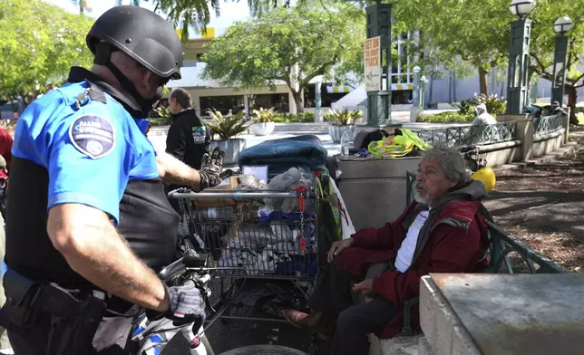 Omar Gonzalez, 70, who is homeless, right, talks with a security guard he sits on a park bench with his belongings as temperatures are forecast to be in the forties this evening Wednesday, Jan. 8, 2025, in Miami. (AP Photo/Lynne Sladky)