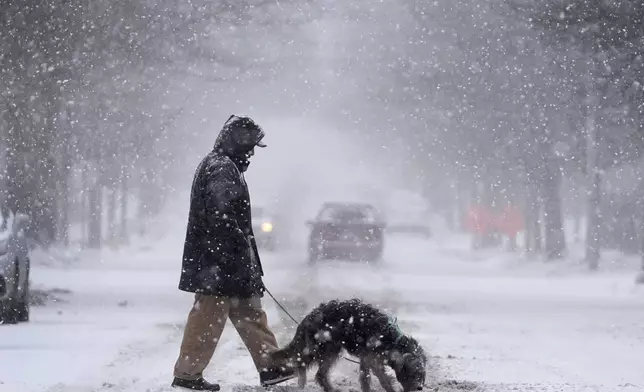 Enrique Davila crosses the street with his dog, Chula, as heavy snow falls Sunday, Jan. 5, 2025, in St. Louis. (AP Photo/Jeff Roberson)