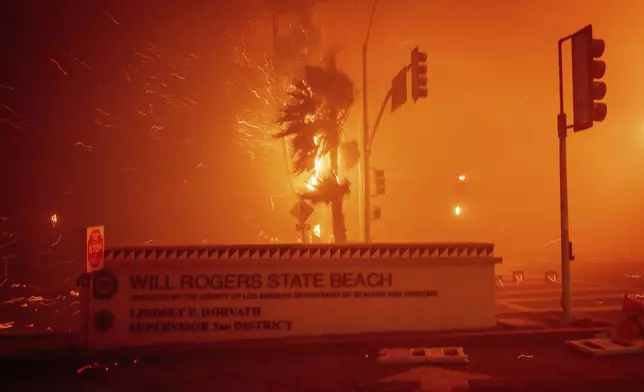 The Palisades Fire burns behind a sign for Will Rogers State Beach in the Pacific Palisades neighborhood of Los Angeles, Tuesday, Jan. 7, 2025. (AP Photo/Ethan Swope)