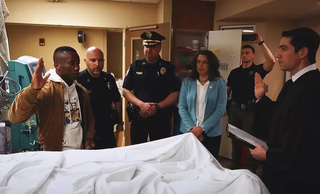 This undated photo provided by the Knoxville, Tenn., Police Department shows Tristants Simpson, a fellow recruit class member, taking the police oath on behalf of Wisbens Antoine, while Sgt. Jimmy Wilson, Police Chief Paul Noel, Mayor Indya Kincannon, Officer Terry Crowe and Municipal Court Judge Tyler Caviness, look on. (Scott Erland/Knoxville Police Department via AP)