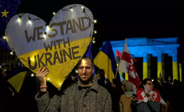 A protester holds a placard at the Brandenburg Gate, illuminated in Ukrainian colors, during a demonstration against Russia's war on Ukraine as they mark the third anniversary of the full-scale invasion in Berlin, Monday, Feb. 24, 2025. (AP Photo/Ebrahim Noroozi)