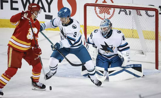 San Jose Sharks' Henry Thrun, center, checks Calgary Flames' Jonathan Huberdeau, left, as Sharks goaltender Alexandar Georgiev, right, looks on during second-period NHL hockey game action in Calgary, Alberta, Sunday, Feb. 23, 2025. (Jeff McIntosh/The Canadian Press via AP)