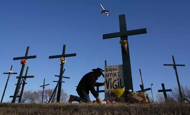 A plane takes off from Ronald Reagan Washington National Airport as Roberto Marquez of Dallas places flowers at a memorial of crosses he erected for the 67 victims of a midair collision between an Army helicopter and an American Airlines jet, Saturday, Feb. 1, 2025, in Arlington, Va. (AP Photo/Carolyn Kaster)