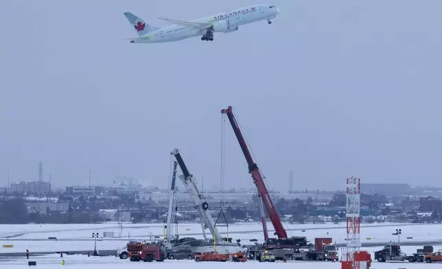 An Air Canada flight takes off in the background as cranes are positioned to remove the wreckage of Delta Flight 4819 from the runway at Toronto Pearson International Airport, in Mississauga, Ontario, on Wednesday, Feb. 19, 2025. (Arlyn McAdorey/The Canadian Press via AP)