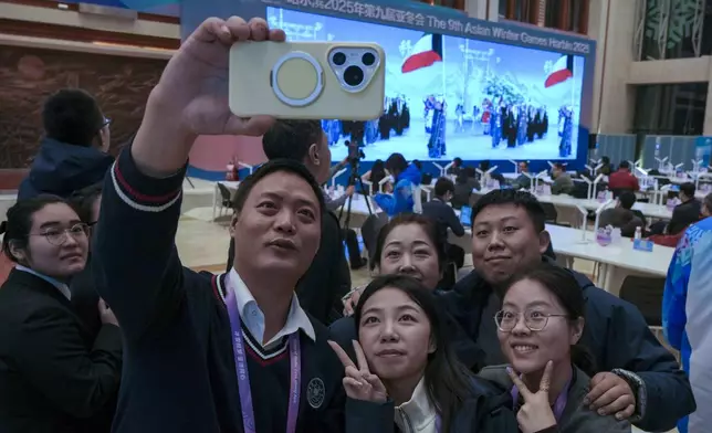 Journalists take a selfie as people watch a screen showing the live telecast of opening ceremony of the 9th Asian Winter Games at the media centre in Yabuli in northeast China's Heilongjiang province on Friday, Feb. 7, 2025. (AP Photo/Andy Wong)