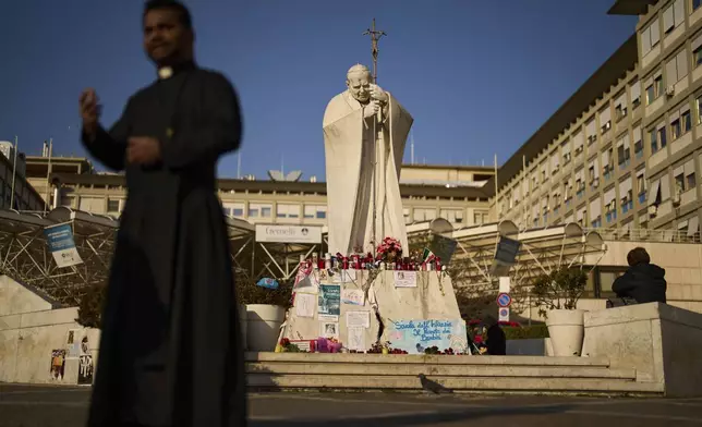 People and a Christian priest stand next to the statue of Pope John Paul II outside the Agostino Gemelli hospital in Rome, Thursday, March 6, 2025. (AP Photo/Francisco Seco)