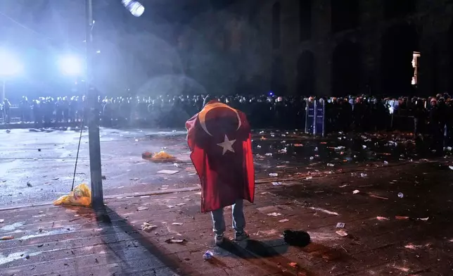A man, a Turkish flag draped on his back, stands in front of anti riot police officers during clashes in a rally against the arrest of Istanbul's Mayor Ekrem Imamoglu, in Istanbul, Turkey, Friday, March 21, 2025. (AP Photo/Khalil Hamra)