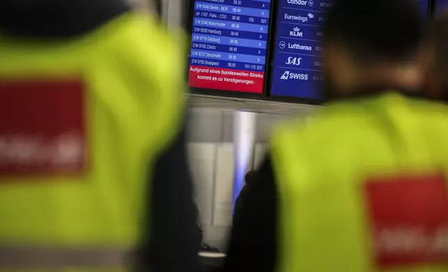 Airport employees wearing high-visibility vests from the Verdi union stand in front of a display board in the departures area at Dusseldorf Airport, Germay with a notice about the strike Monday, March 10, 2025. (Christoph Reichwein/dpa via AP)