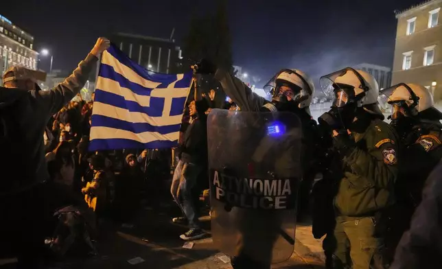 Protesters hold a Greek flag next to riot police during clashes, on the third and final day of a censure motion debate against the conservative government in parliament over a deadly rail disaster nearly two years ago, in Athens, Greece, Friday, March 7, 2025. (AP Photo/Petros Giannakouris)