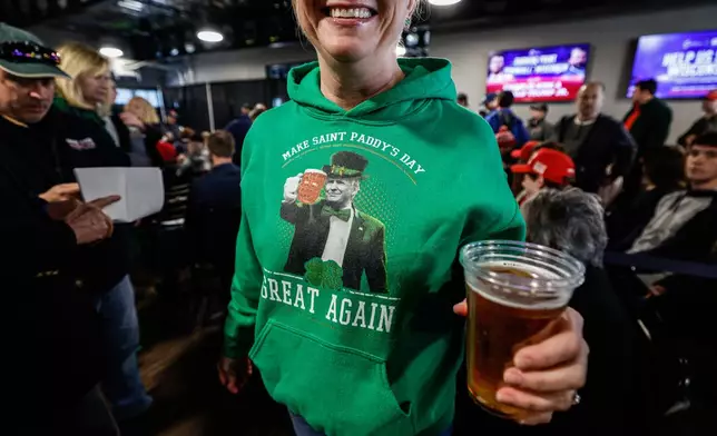 A woman wearing a Donald Trump and Saint Patrick's Day themed hoodie enjoys a beer before a town hall meeting Monday, March 17, 2025, in Oconomowoc, Wis. (AP Photo/Jeffrey Phelps)