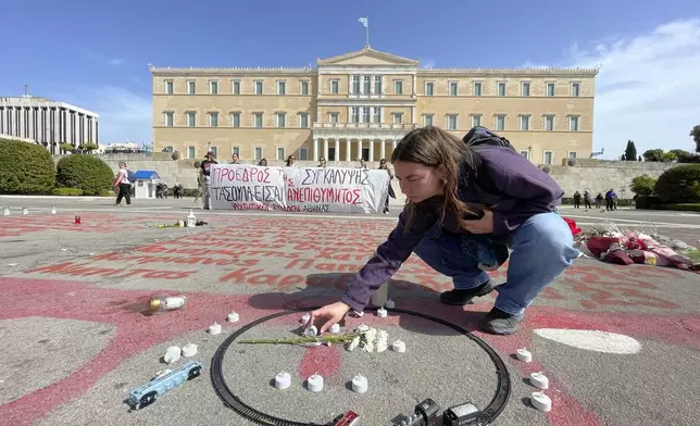 University student Despoina Billia places candles on the ground next to a toy train set at a makeshift memorial for a deadly 2023 rail disaster, outside parliament where Greece's new President Constantine Tassoulas was sworn in, in Athens, Greece, Thursday, March 13, 2025. The names of the victims are written in red paint on the ground, while the banner in the background reads "Coverup President - Tassoulas Unwanted." (AP Photo/Lefteris Pitarakis)