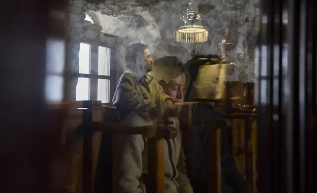 Newly married French couple Thomas and Valentine attend the Divine Liturgy as Constantin Papakonstantinou, an assistant residing at the monastery, left, chants as he is reflected on a window inside the chapel of the Monastery of Panagia Hozoviotissa in Amorgos island, Greece, on Tuesday, March 25, 2025. (AP Photo/Petros Giannakouris)